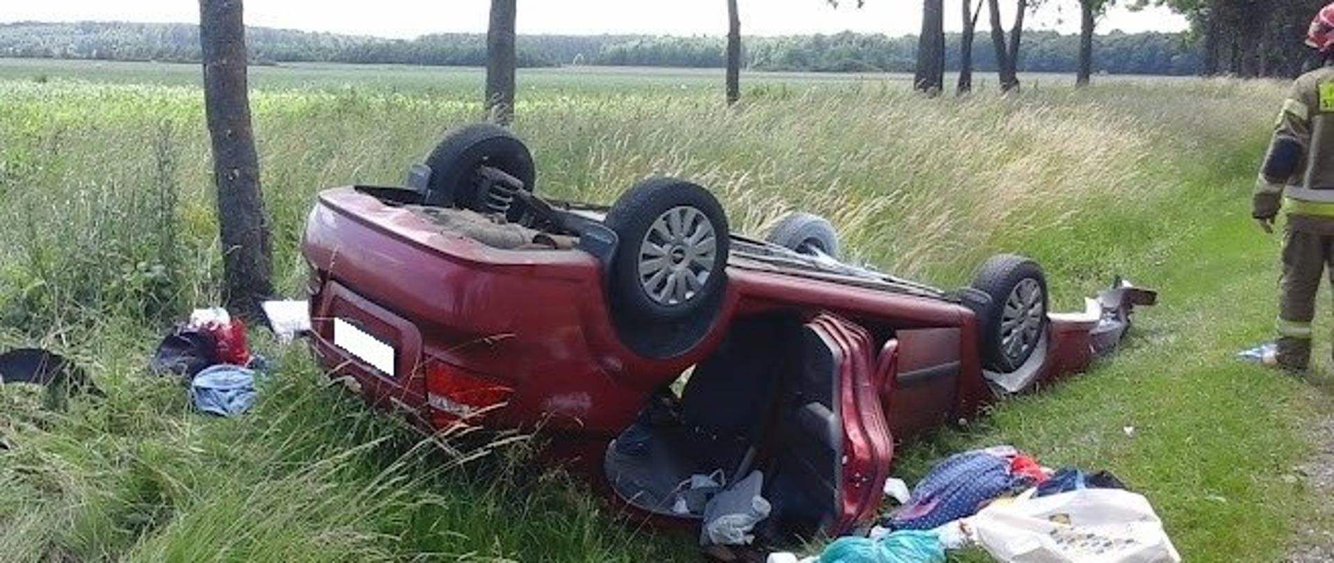 Zdjęcie przedstawia samochód osobowy koloru czerwonego lezący na dachu w przydrożnym rowie, obok rozbitego pojazdu leżą rzeczy podróżujących