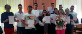Zdjęcie grupowe: grupa uczniów i nauczycielka w różowych spodniach trzymają świadectwa i kwiaty.