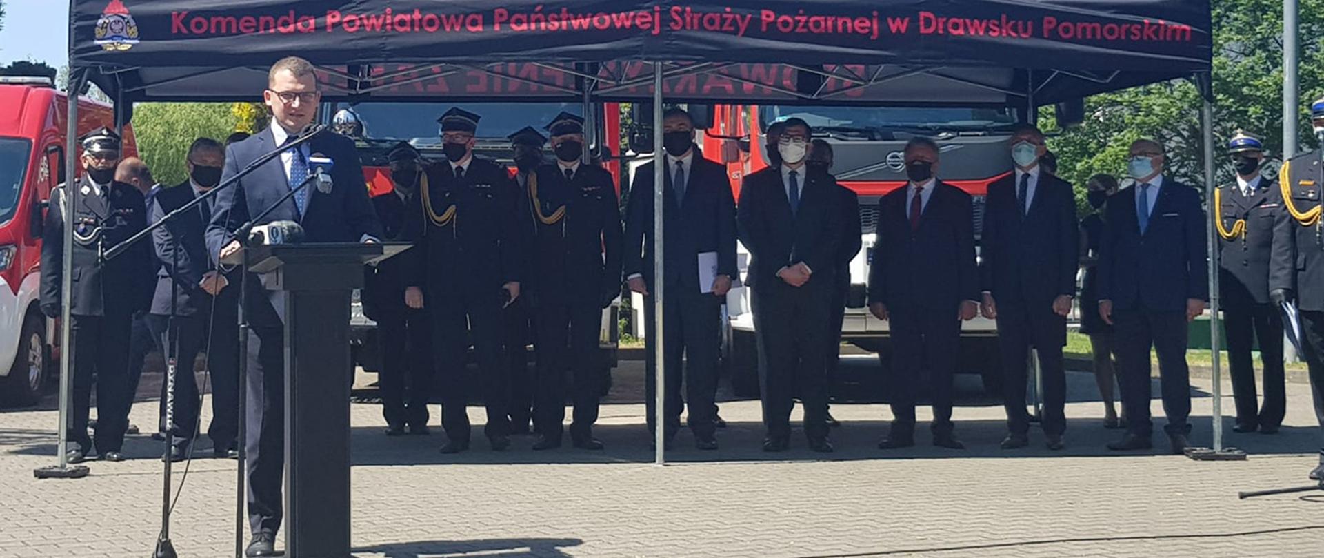 Na zdjęciu widać Pawła Szefernakera wiceministra MSWiA stojącego przy mównicy i przemawiającego. W tle widać innych uczestników uroczystości stojących pod dużym namiotem z napisem " Komenda Powiatowa Państwowej Straży Pożarnej w Drawsku Pomorskim.