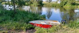 Na zdjęciu widać łódź wiosłową, czerwoną. Łódź zwodowana. Kolor czerwony. Na drugim planie rzeka, kolor wody czarny. Niebo niebieskie. Zdjęcie w ciągu dnia. Brzegi porośnięte zieloną roślinnością.