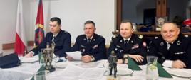 umundurowani w mundurach wyjściowych czterech strażaków, wśród nich zastępca komendanta głównego nadbryg. Adam Konieczny, siedzący przy stole w obiekcie świetlicy, w lewym narożniku widoczne flagi