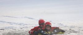Dwóch ratowników w czerwonych kombinezonach i kaskach ochronnych realizuje symulację akcji ratunkowej na zamarzniętym akwenie. Częściowo zanurzeni w lodowatej wodzie, wykorzystują żółty sprzęt wypornościowy oraz czerwoną linę asekuracyjną, zapewniającą bezpieczeństwo i kontrolę nad przebiegiem działań. Wokół nich widoczne są pokrywa śnieżna i połamany lód, co podkreśla realistyczne warunki terenowe. Ćwiczenie ma na celu doskonalenie technik ratownictwa wodnego w ekstremalnych warunkach zimowych oraz sprawdzenie skuteczności procedur i sprzętu w sytuacji zagrożenia życia.