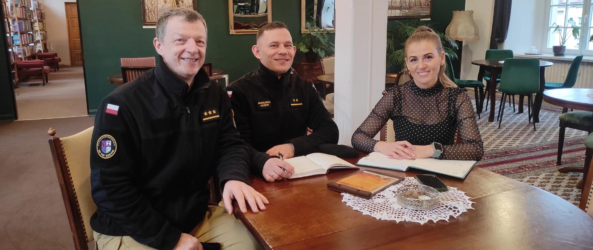 Zdjęcie przedstawia spotkanie w zamkowej kawiarni biblioteki. Przy stole znajdują się trzy osoby, dwóch strażaków w czarnych mundurach oraz kobieta u brana na czarno.