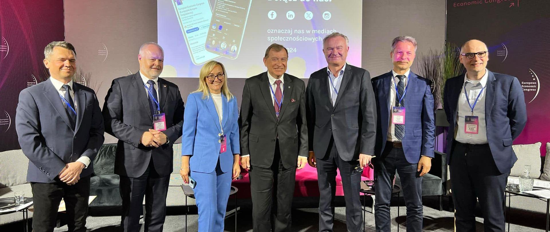 Pod fioletową ścianką z napisem Dołącz do nas stoi w szeregu siedem osób, drugi z lewej wiceminister Gzik.