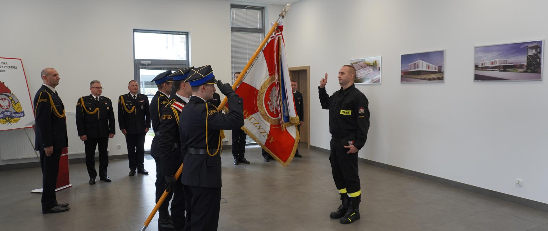 Zdjęcie przedstawia ślubowanie nowo przyjętego funkcjonariusza Komendy Miejskiej PSP w Rzeszowie w umundurowaniu koszarowym z ręką uniesioną w górze na wprost pocztu sztandarowego Komendy Miejskiej PSP w Rzeszowie. W tle widoczni są funkcjonariusze w mundurach wyjściowych.