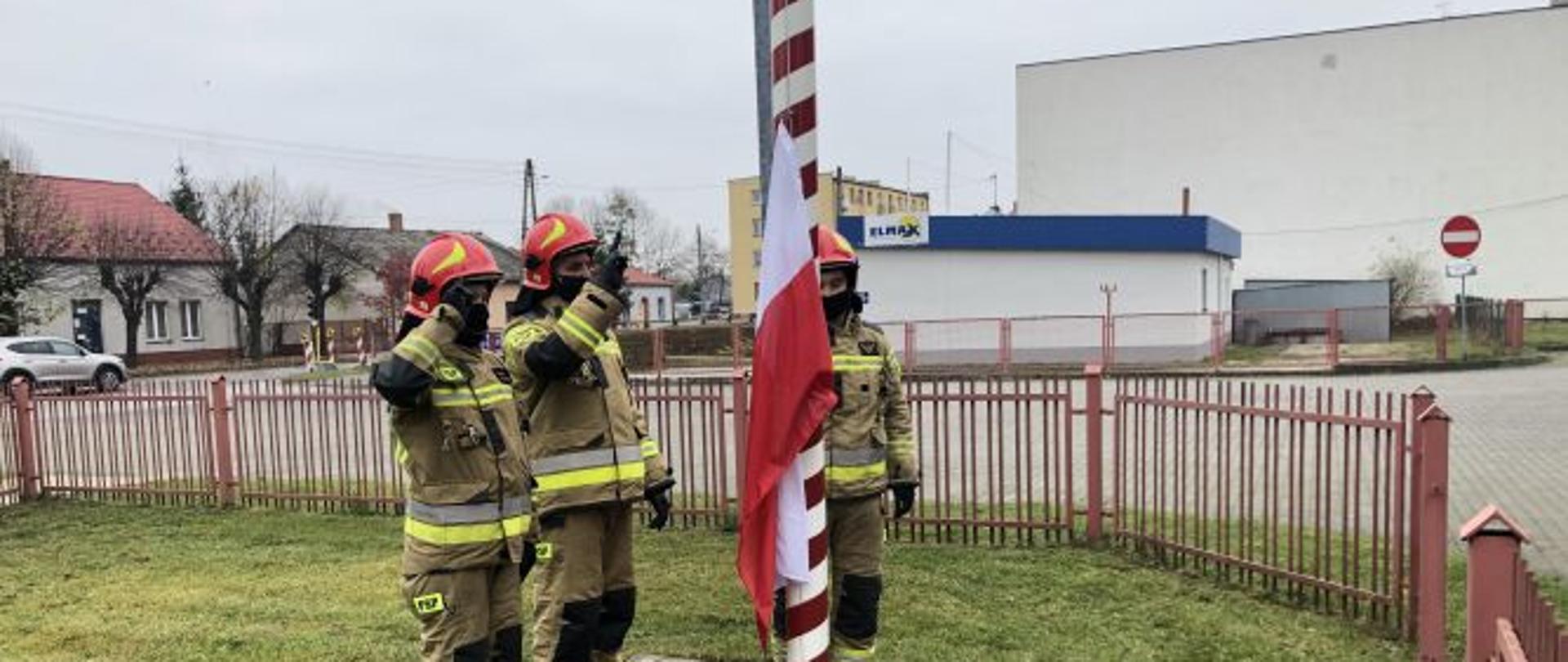 zdjęcie przedstawia salutujących trzech strażaków -poczet flagowy - przy maszcie na którym zawieszona jest flaga. zdjęcie wykonano z boku masztu. w tle widać budynki mieszkale i wybrukowany plac zewnętrzny komendy. 