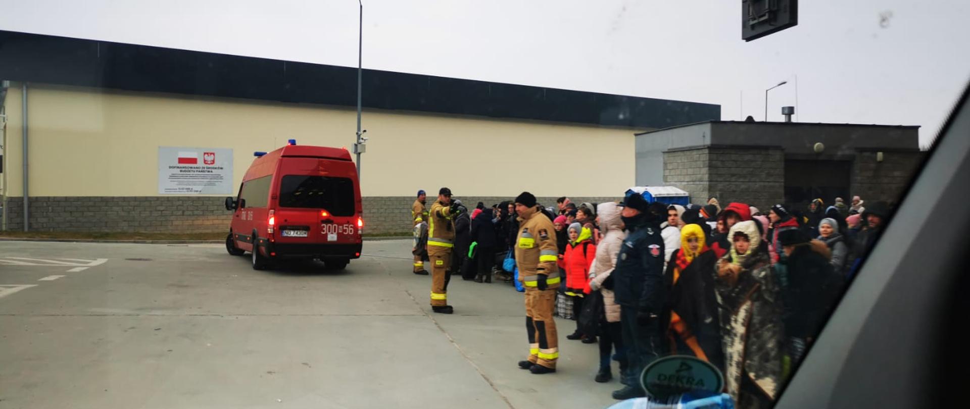 Zdjęcie wykonane zza szyby pojazdu. Na fotografii widoczny plac zamknięty ścianą budynku z umieszczoną na niej tablicą z flagą i godłem Rzeczypospolitej. Po prawej stronie stoją w dużej grupie zgromadzeni ludzie w ciepłych okryciach, niektórzy w kapturach i owinięci kocami. Od placu odgradza ich trzech strażaków w piaskowych mundurach bojowych i ciemnych czapkach na głowach. Na końcu placu widać tył odjeżdżającego czerwonego strażackiego mikrobusa.