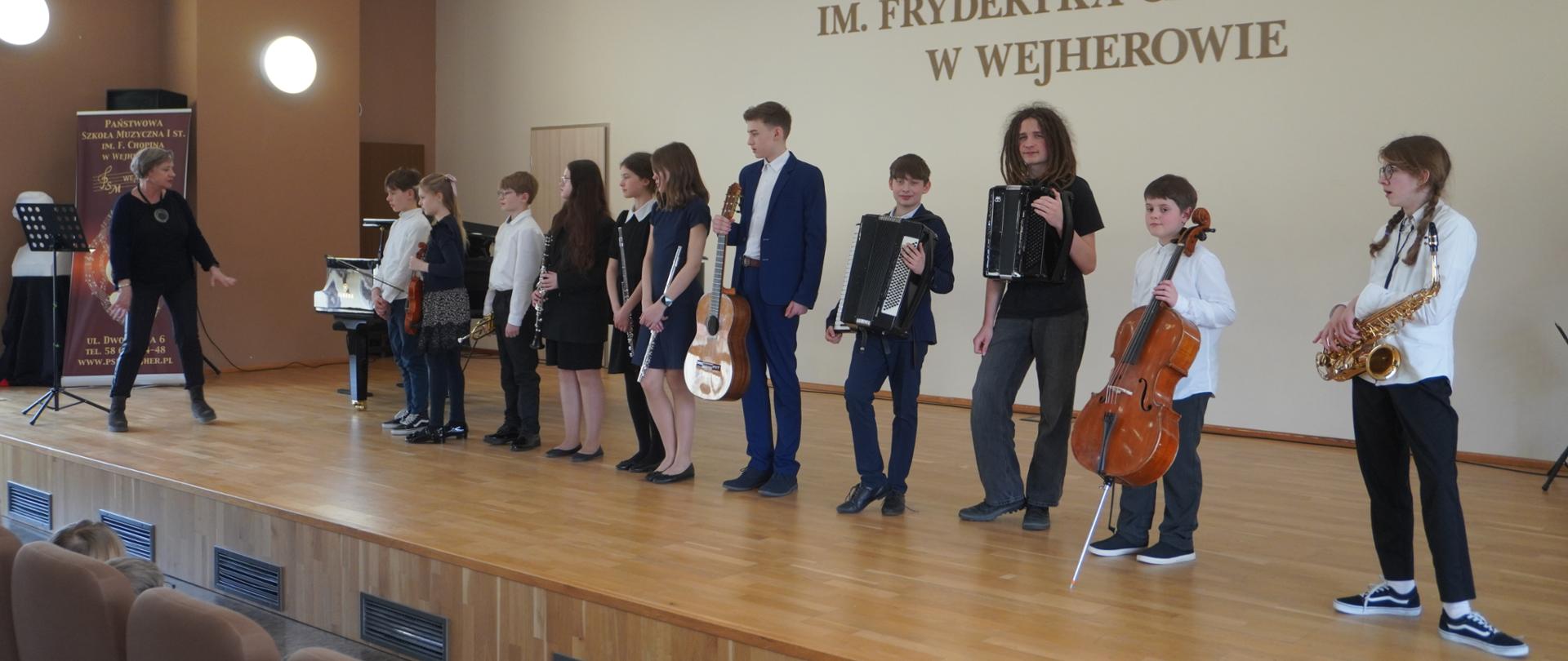 Na estradzie uczniowie występujący podczas koncertu prezentują swoje instrumenty. Stoją i eksponują je młodej publiczności