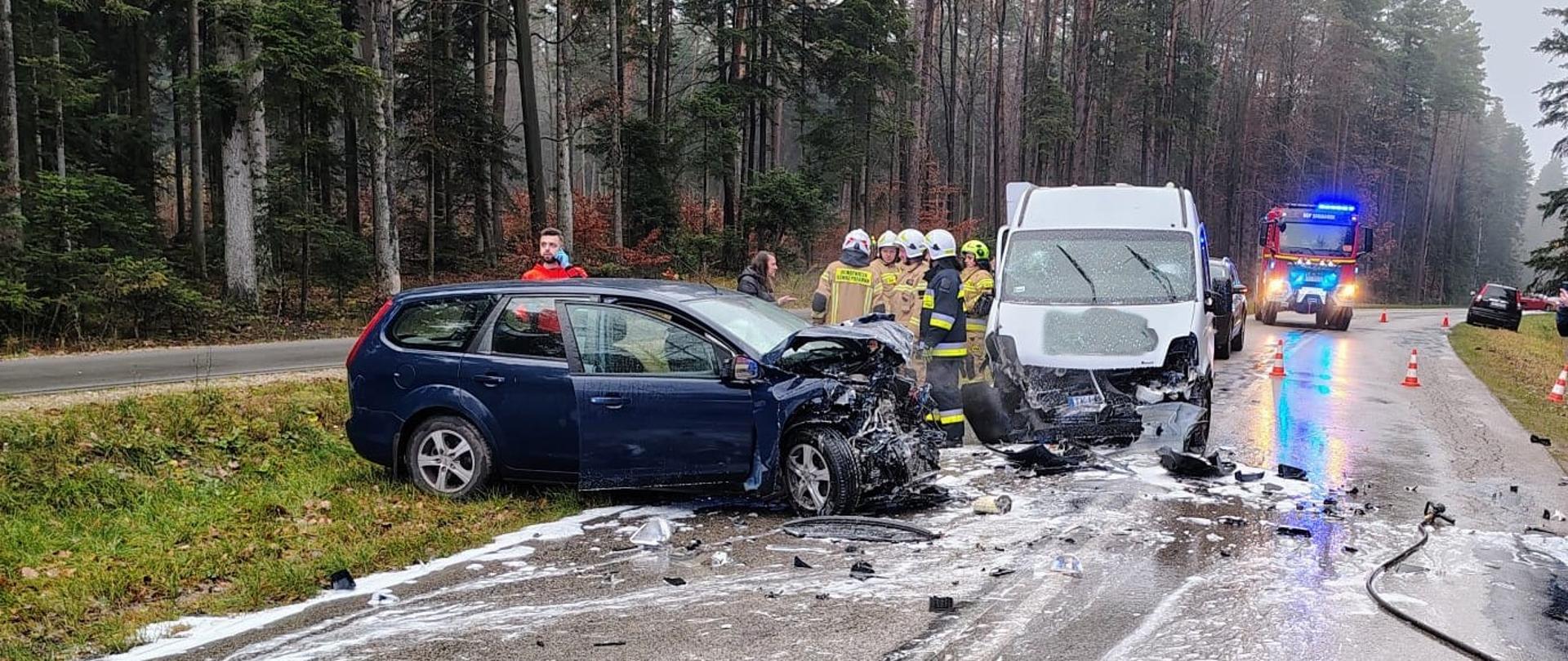 Zdjęcie przedstawia miejsce wypadku:. Na jezdni stoją dwa uszkodzone samochody, przy nich grupa ratowników, w głębi widać samochód pożarniczy. 