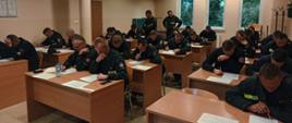 Widok z przodu. Sala wykładowa. Słuchacze szkolenia pochyleni nad biurkami w trakcie egzaminu teoretycznego. 