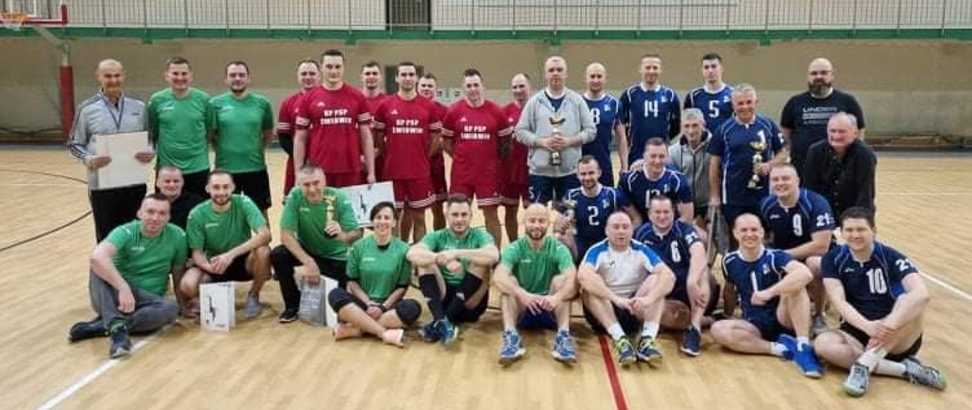 Zdjęcie przedstawia cztery drużyny pozujące do zdjęcia po turnieju piłki siatkowej na hali sportowej. Mężczyźni na zdjęciu ubrani są w koszulki: zielone, czerwone, niebieski i czarne co odpowiada przynależności do danej drużyny. Kliku z nich trzyma w ręku dyplomy i puchary.