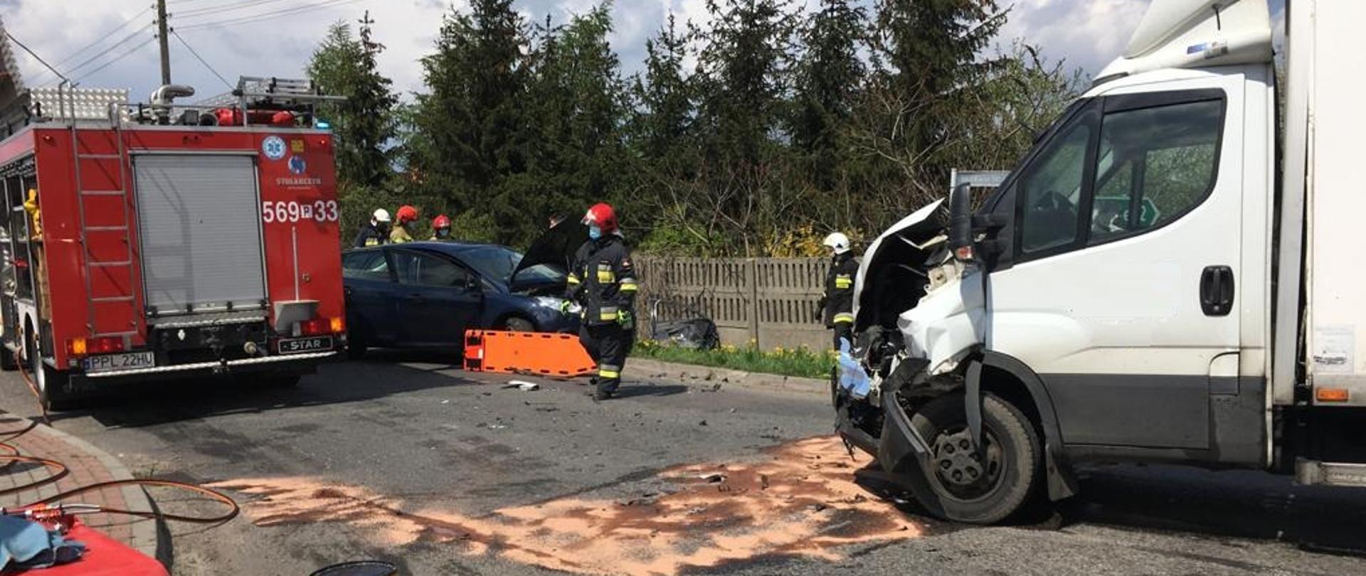 Zdjęcie zrobione w czasie kolizji drogowej. Na środku drogi stoi samochód dostawczy, który ma uszkodzony przód auta. Przed nim za pomocą środka sorpcyjnego został zneutralizowany wyciek substancji ropopochodnych. Na zdjęciu widoczny jest tył samochodu strażackiego. Po lewej stronie leżą szczotki i narzędzia hydrauliczne. 