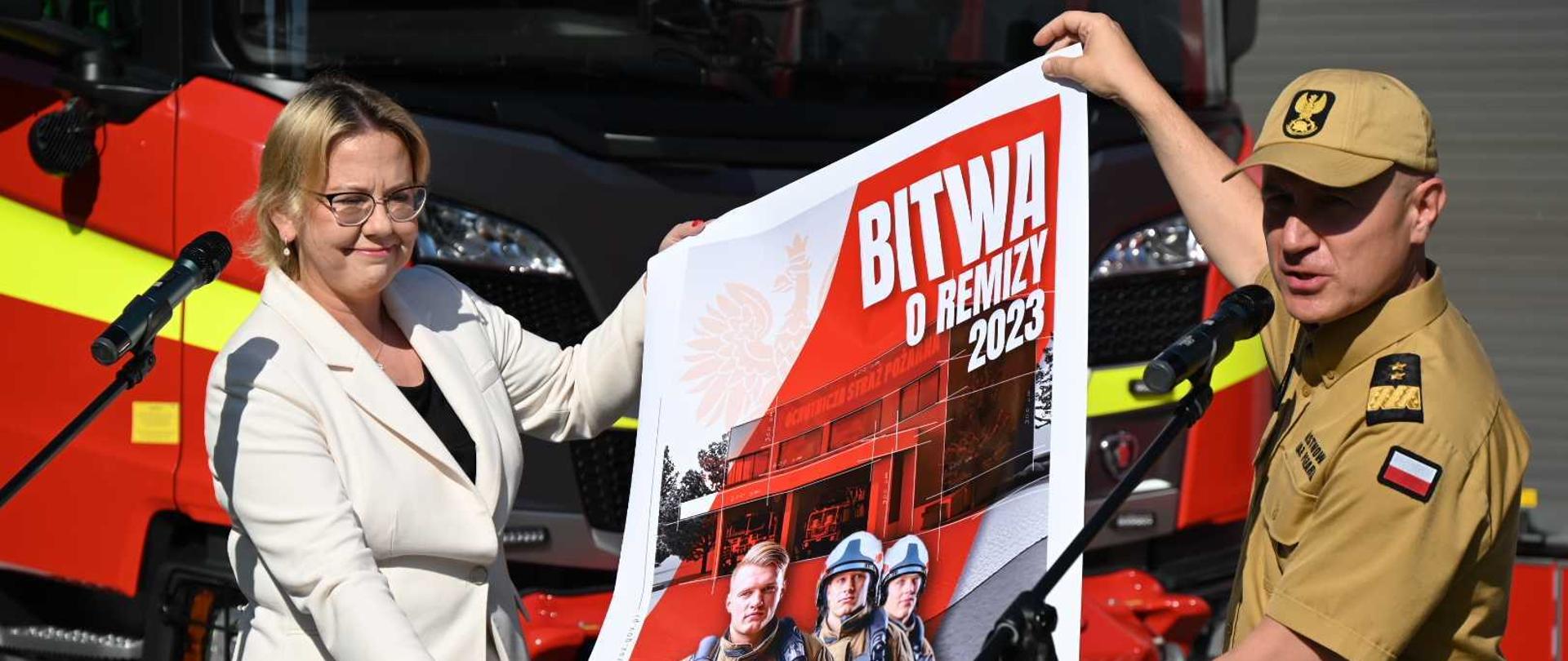 Komendant główny psp generał brygadier andrzej bartkowiak oraz Anna moskwa Minister klimatu i środowiska Polski przedstawiają plakat z nową akcją bitwa o remizy , oboje stoją przed mikrofonem na tle wozu bojowego