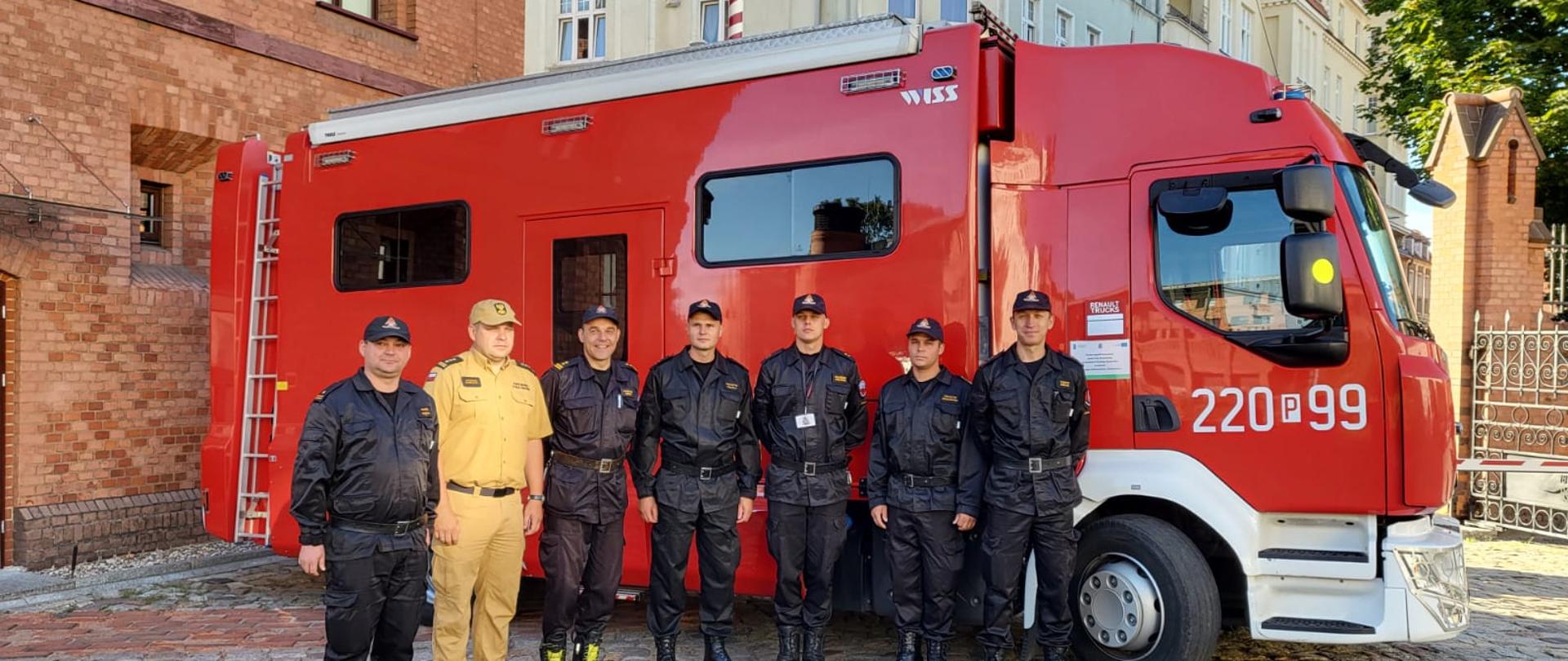 grupqa strażaków stoi w szeregu, za nimi duże czerwone auto ora budynek z cegły