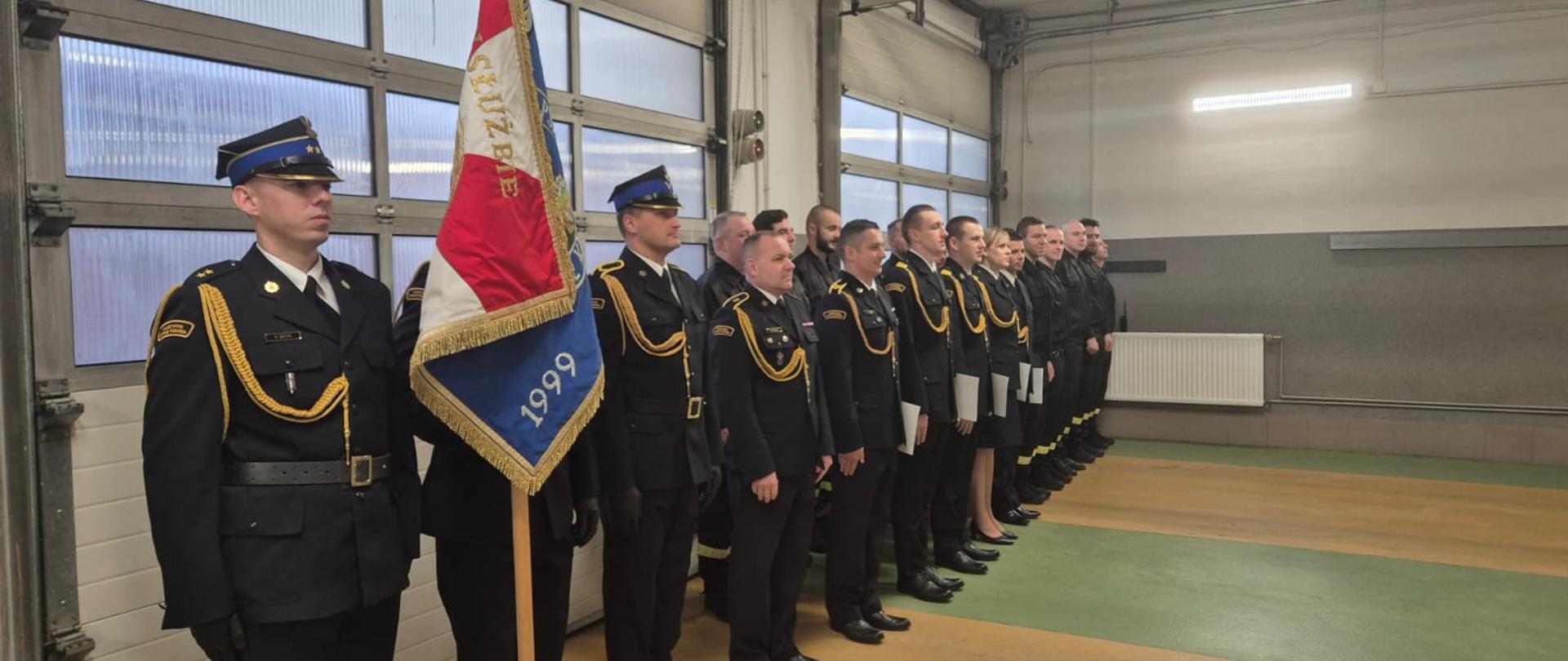 Strażacy zgromadzeni w garażu budynku Komendy powiatowej państwowej straży pożarnej w sępólnie krajeńskim w mundurach wyjściowych i koszarowych w obecności komendanta powiatowego podczas uroczystości święta niepodległości