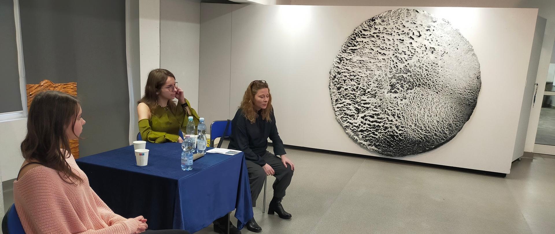 W galerii szkolnej Magdalena Soboń siedzi obok stoika przykrytego granatowym materiałem. Kobieta jest w średnim wieku. Ma długie włosy. Ubrana jest w ciemne spodnie i czarny sweter. Obok stolika siedzą dwie uczennice. Na stoliku znajdują się butelki z wodą, kartonowe kubki i zadrukowana kartka. W tle na ściance działowej wisi duża praca artystki. Jest to abstrakcja. Przypomina czarnobiałą kulę ziemską z chropowatą powierzchnią