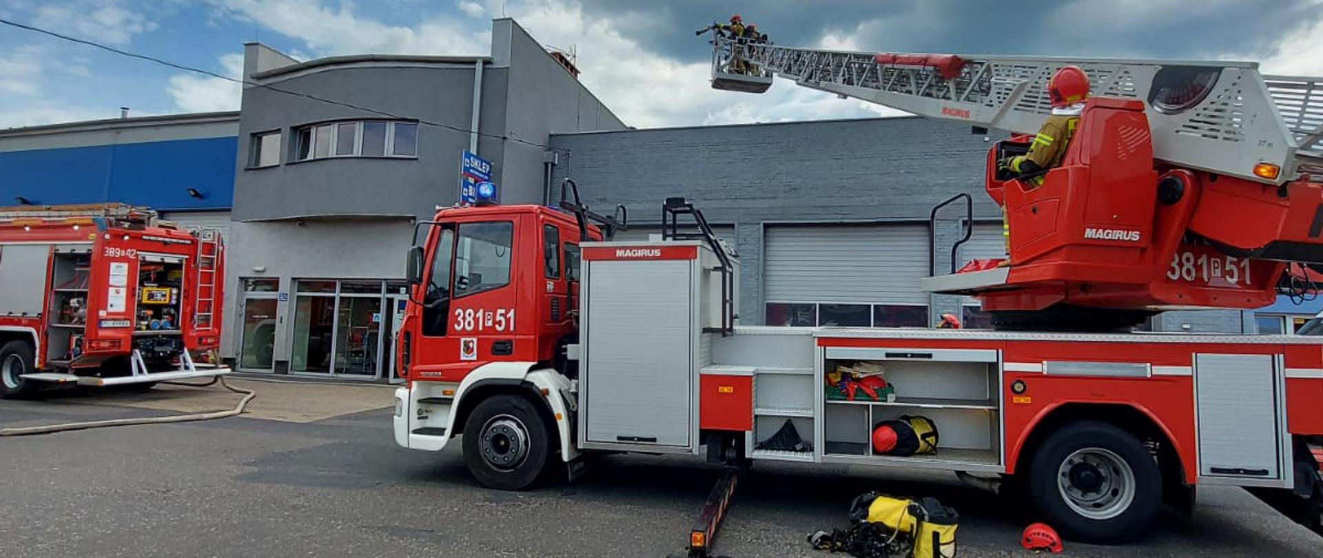 Dwa samochody pożarnicze na placu przed budynkiem warsztatów. Z lewej samochód gaśniczy z rozwinięta linią gaśniczą, z prawej drabina mechaniczna wysunięta ponad dach budynku. Strażacy w koszu na drabinie.