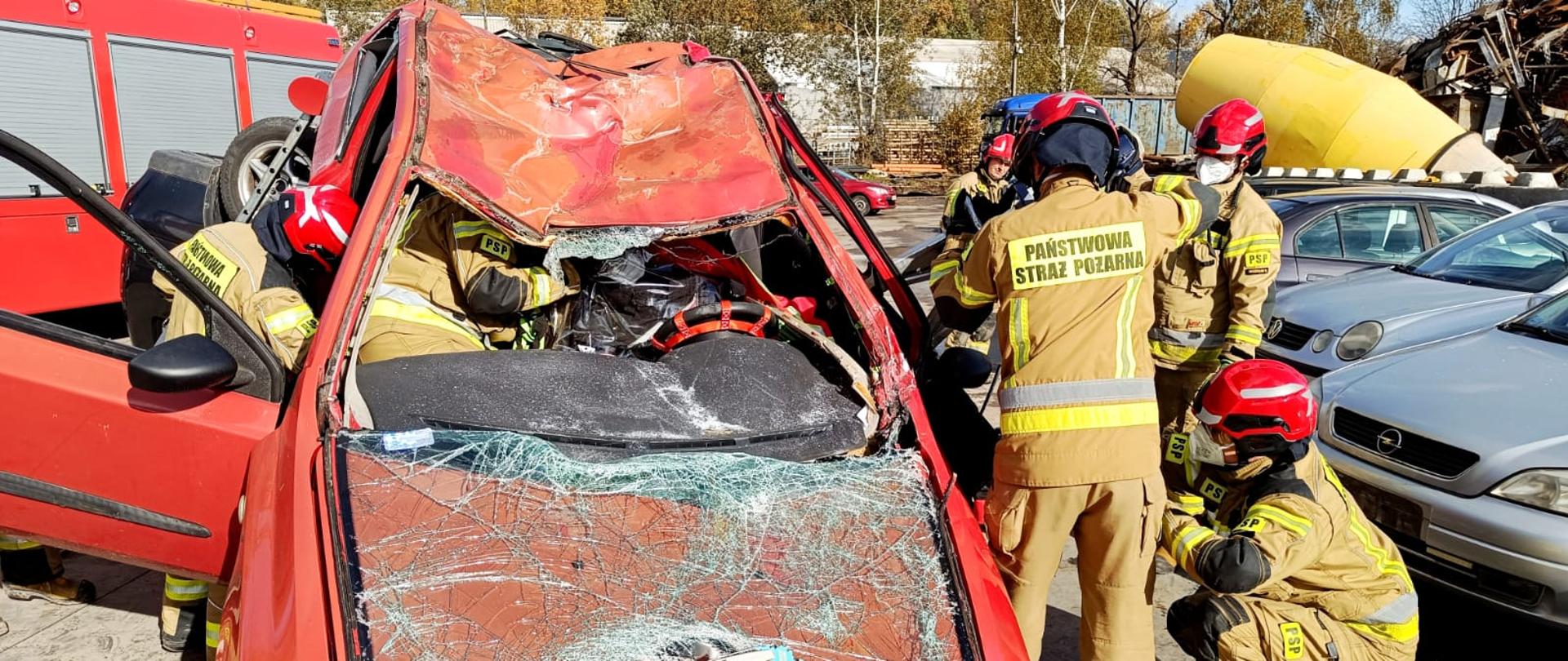 Strażacy z Państwowej Straży Pożarnej w trakcie ćwiczeń ratowniczych przy rozbitym czerwonym samochodzie marki Fiat. Pojazd ma poważnie zgnieciony dach i rozbitą przednią szybę. Ratownicy w hełmach i ubraniach ochronnych wykorzystują sprzęt hydrauliczny do wydobycia osoby z wnętrza auta. W tle widoczne są inne pojazdy oraz wóz strażacki.