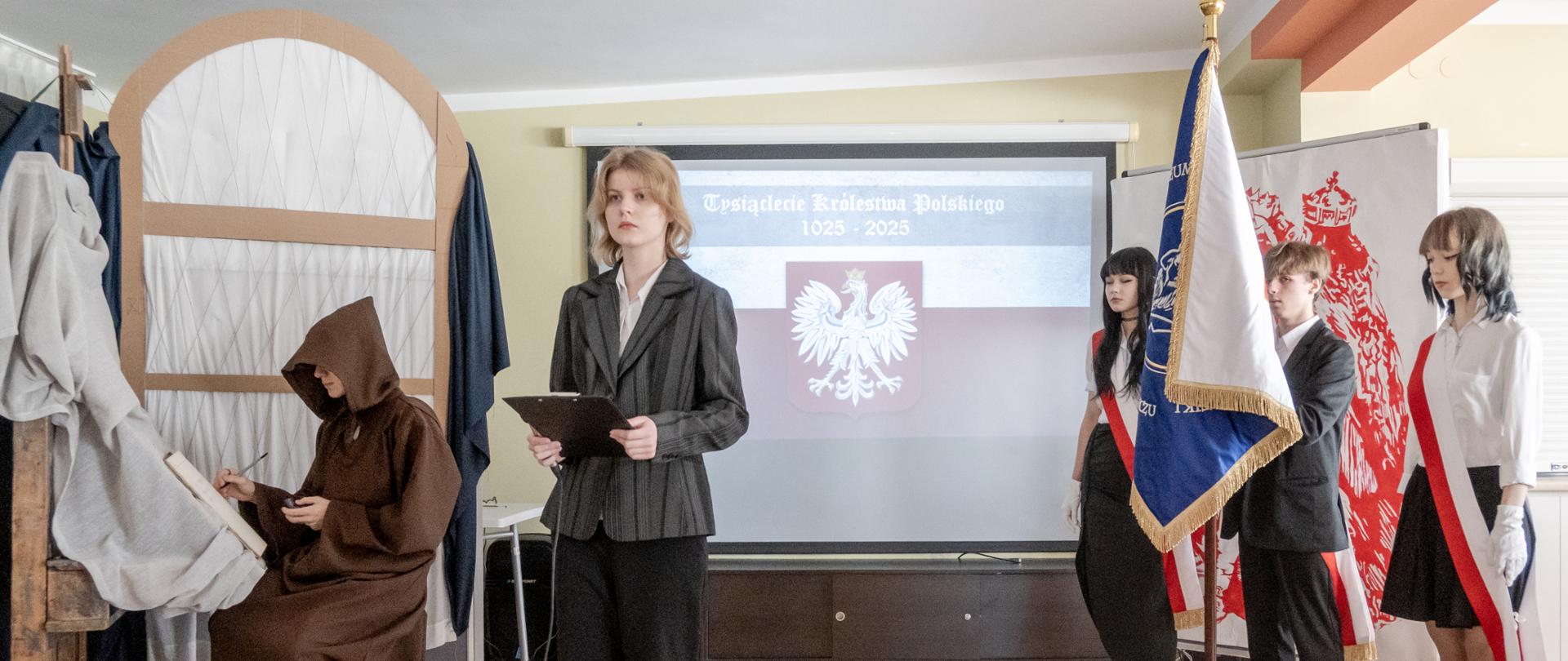W szkolnej sali odbywa się uroczystość historyczno-artystyczna. Na pierwszym planie stoi uczennica w garniturze z mikrofonem i czarnym folderem. Po lewej stronie sceny siedzi uczeń przebrany za średniowiecznego mnicha, tworzący iluminowany manuskrypt przy stylizowanym pulpicie. W tle, na ekranie, widoczny jest slajd z napisem „Tysiąclecie Królestwa Polskiego 1025–2025” i herbem Polski. Po prawej stronie uczniowie w galowych strojach z biało-czerwonymi szarfami trzymają sztandar szkoły. W tle dekoracja z czerwonym wizerunkiem Bolesława Chrobrego.