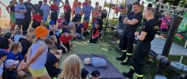 Zdjęcie przedstawia funkcjonariuszy KP PSP podczas wizyty na obozach MDP w Wieleniu. Na zdjęciu widać grupę dzieci w mundurkach strażackich oraz przedstawicieli PSP prowadzących pokaz udzielania pierwszej pomocy. Zdjęcie wykonane na zewnątrz, ładna pogoda.