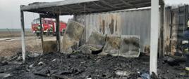 Na zdjęciu widzimy spalony kontener, zgliszcza. Część blaszanych płyt oparta jest o drugi kontener. Z lewej strony kadru widzimy w oddali samochód strażacki koloru czerwonego oraz dwóch strażaków ubranych w odzież specjalną koloru czarnego, na głowie białe hełmy.