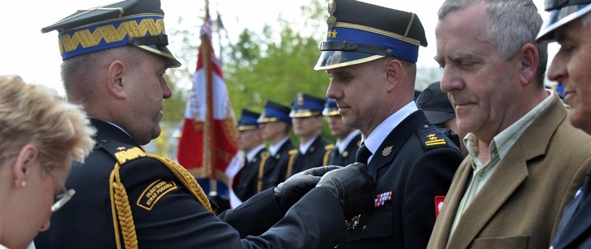 Zdjęcie zrobione na zewnątrz podczas uroczystości. Funkcjonariusz Państwowej Straży Pożarnej w stopniu generała wręcza odznaczenie strażakowi, który jest ustawiony w szeregu razem z innymi osobami odbierającymi odznaczenia. 
