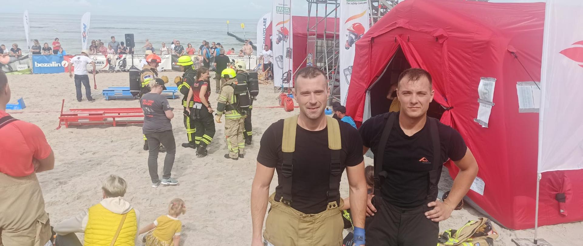 Zdjęcie przedstawia dwóch strażaków spodniach bokowych i krótkich koszulkach stojących obok siebie na plaży. W tle czerwone namioty, metalowa wieża, inni ludzie i morze.