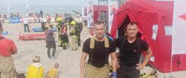 Zdjęcie przedstawia dwóch strażaków spodniach bokowych i krótkich koszulkach stojących obok siebie na plaży. W tle czerwone namioty, metalowa wieża, inni ludzie i morze.