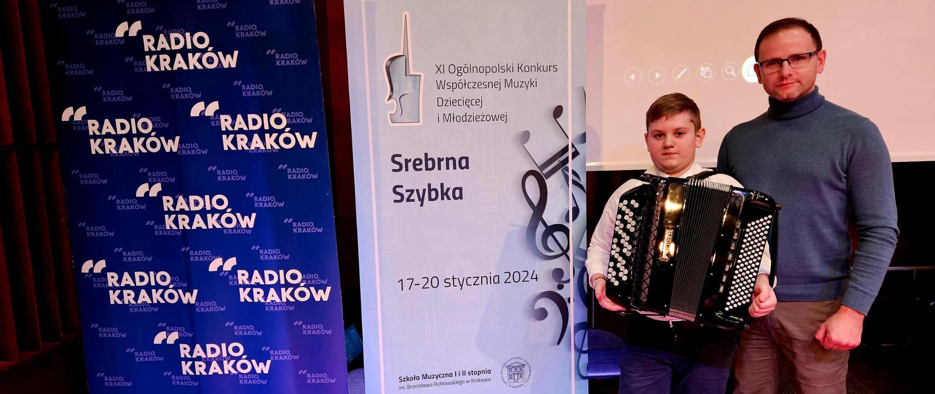 Zdjęcie nauczyciela mgra Krzysztofa Burego oraz jego ucznia Szymona Kukli obok rollupów reklamujących konkurs oraz Radio Kraków
