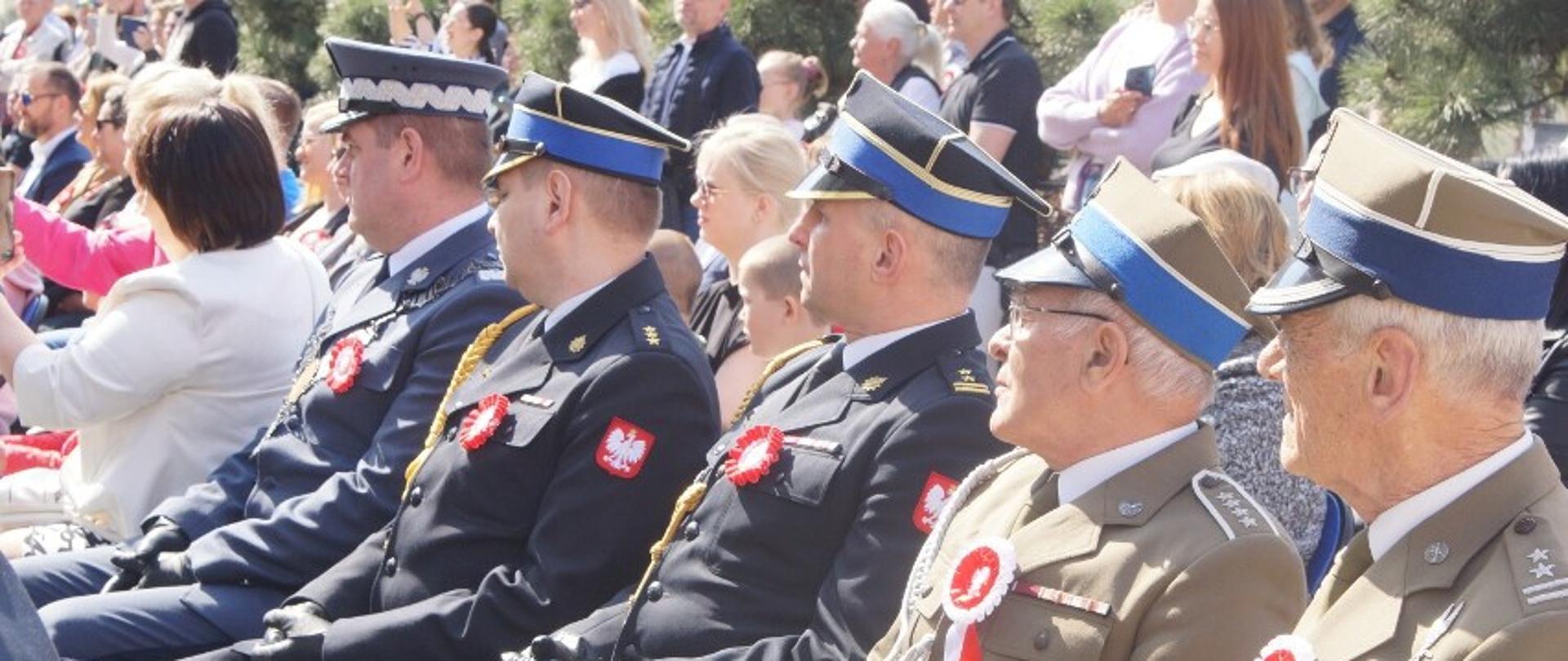 Na zdjęciu widać osoby w mundurach służbowych. Zdjęcie wykonane na rynku, przed ratuszem. Zdjęcie wykonane w ciągu dnia. Osoby mają przypięte białoczerwone kotyliony do munduru.