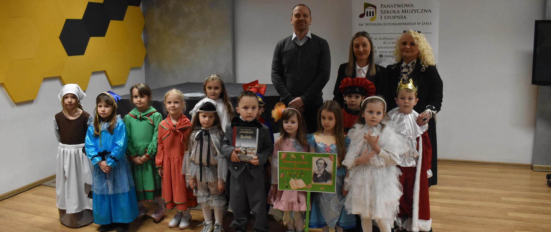 Na zdjęciu, w sali kameralnej PSM I st. w Jaśle stoją w szeregu wychowankowie Bajkowego Przedszkola w Jaśle poprzebierani w stroje z różnych bajek Andersena, a także wychowawcy Bajkowego Przedszkola w Jaśle oraz nauczyciel PSM I st. w Jaśle. Kolorystyka zdjęcia jest biało-szaro-żółto-czarno-czerwono-niebiesko-zielona. 