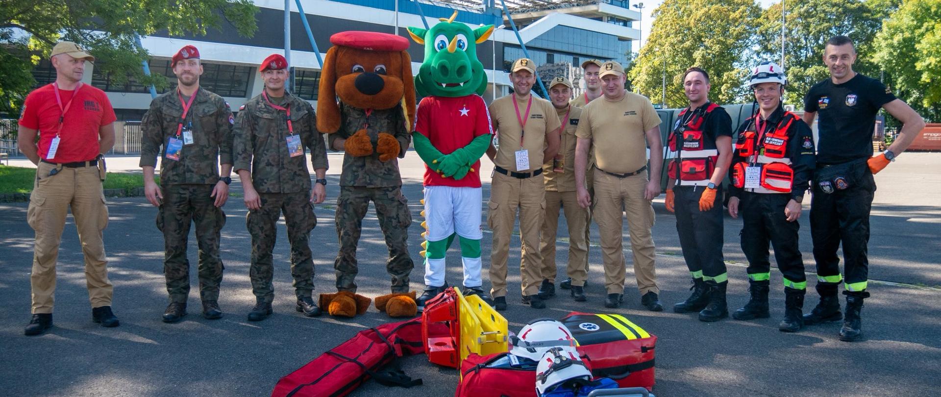 Grupa uczestników zawodów, w tym strażacy PSP, żołnierze oraz maskotki, stojąca wspólnie na placu przed stadionem w Krakowie. Na pierwszym planie widoczne torby medyczne i kaski ratownicze.
fot. Michał Mizia