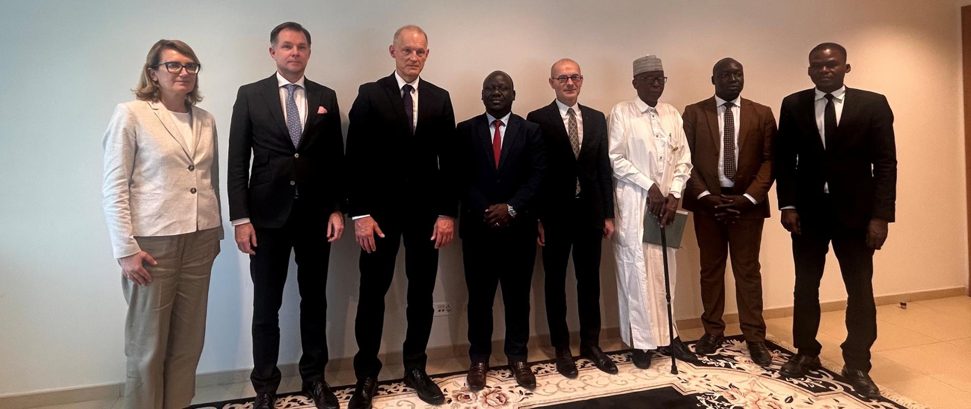 A group of nine people (one woman, eight men) poses for a group photo during a diplomatic meeting; the participants, dressed in formal attire—suits, with one person wearing traditional white clothing—stand in a room against the backdrop of a light-colored wall. Deputy Minister Zajączkowski is pictured (third from the left).