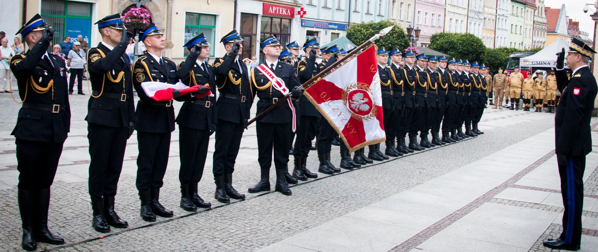 Rynek, strażacy w umundurowaniu galowym stoją w szeregu, sztandar, przed generałem
