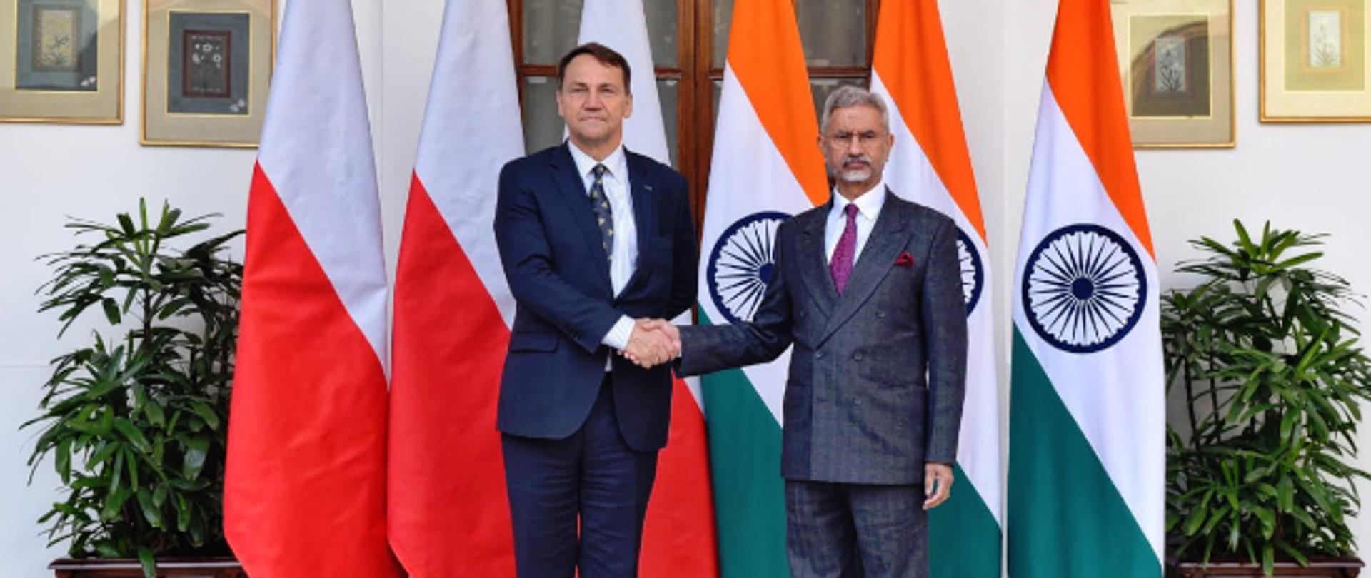 Two men wearing suits stand facing each other and shake hands during an official meeting. Polish and Indian flags are displayed behind them in a representative interior.