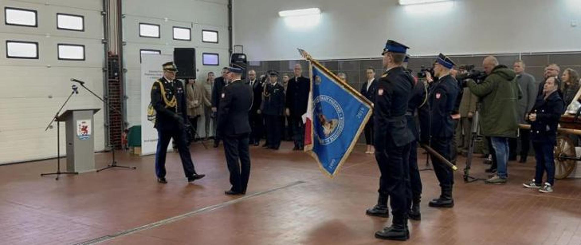 Zdjęcie przedstawia moment przejęcia obowiązków przez Komendanta. W tle widać zgromadzonych oficjeli. Przed Komendantami poczet flagowy Komendy PSP w Białogardzie.