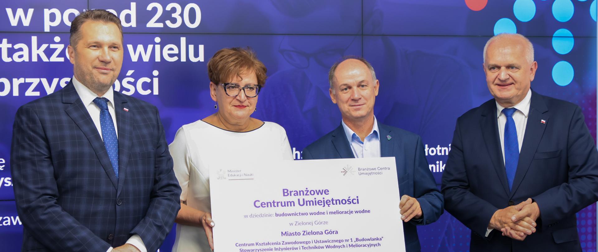 Na tle dużego ekranu stoi trzech mężczyzn i kobieta. Kobieta z mężczyzną trzyma dużą plansze z napisem Branżowe Centrum Umiejętności 10 mln zł. 