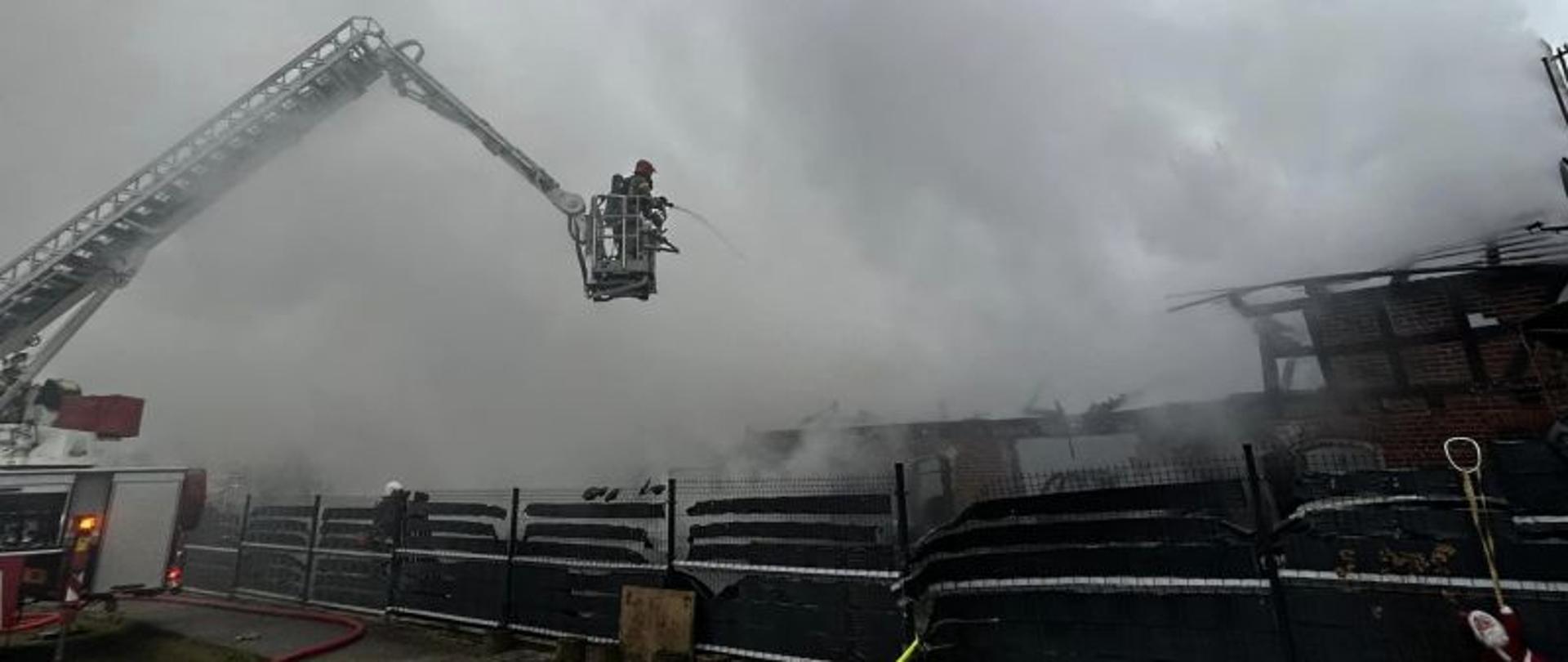 Centralny punkt zdjęcia stanowi ramię podnośnika. Tło stanowi palący się budynek. Nie widać ognia ale bardzo duże zadymienie. W koszu podnośnika strażacy.