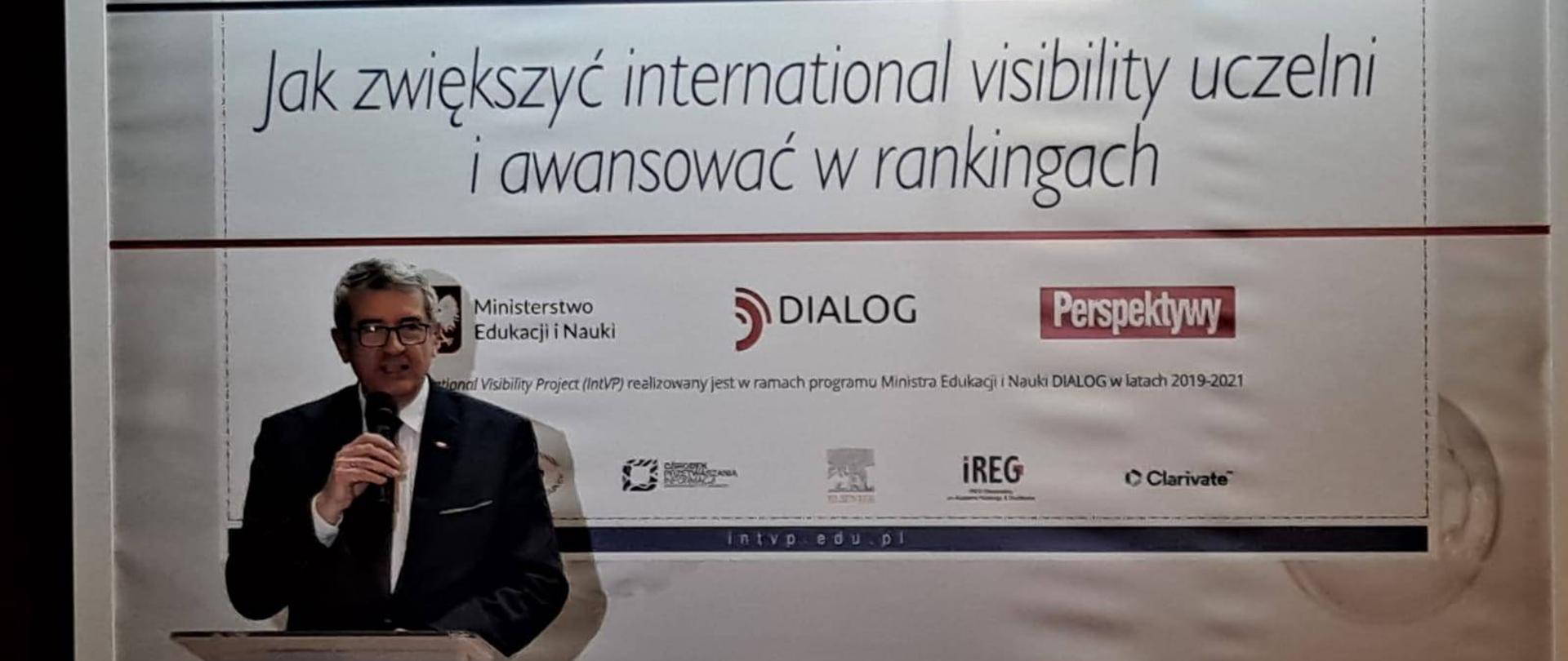 Perspektywy – seminarium z udziałem wiceministra Wojciecha Murdzka. wiceminister przemawia do mikrofonu przy mównicy. W tle plansza z napisem jak zwiększyć widoczność uczelni i awansować w rankingach 