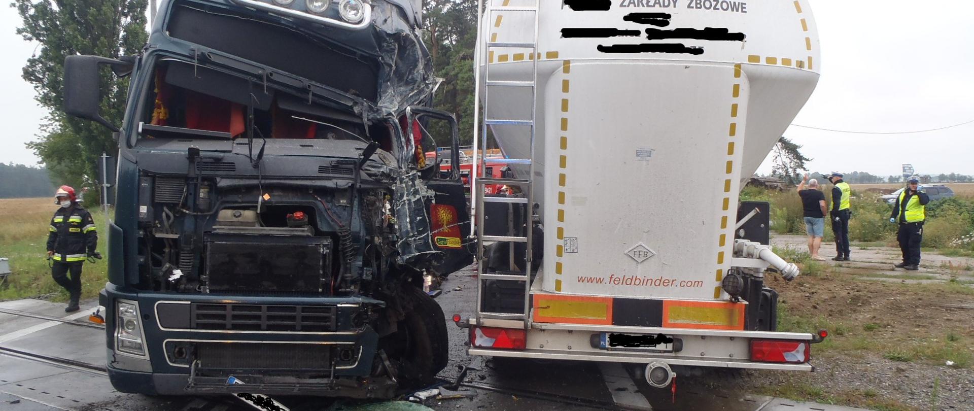 dwie rozbite ciężarówki stoją na torowisku, jedna przodem z rozbitą kabiną druga tyłem, w tle strażak i policjanci