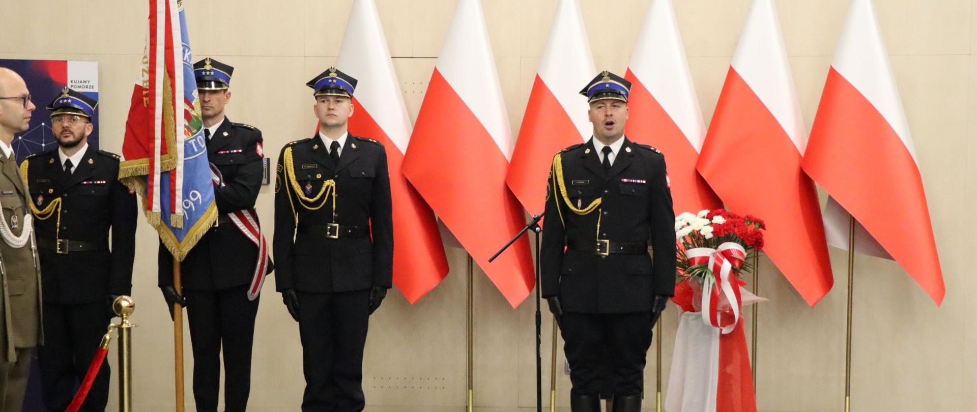 Widok na dowódcę uroczystości, który śpiewa. Stoi w pozycji zasadniczej. Z lewej strony poczet sztandarowy strażaków w umundurowaniu reprezentacyjnym. Za nimi widać mównicę, w tle dekoracja z biało-czerwonych flag.