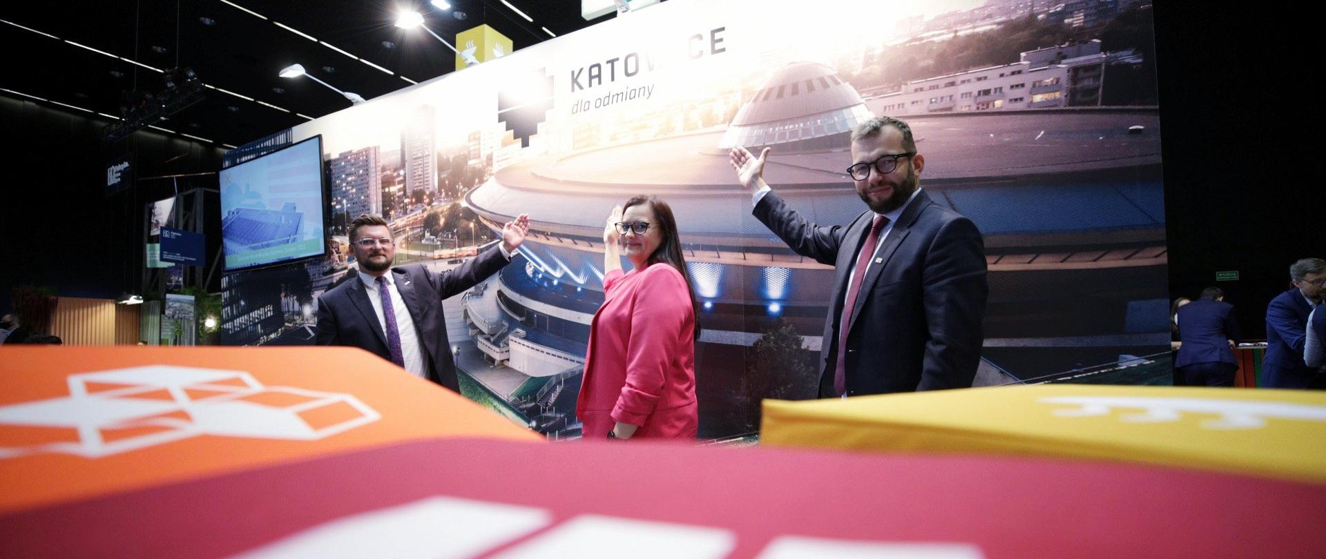 Minister Małgorzata Jarosińska-Jedynak, wiceminister Grzegorz Puda i prezydent Katowice Marcin Krupa podczas EKG 2020, stoją na tle zdjęcia przedstawiającego spodek, z przodu kolorowe elementy przestrzenne.