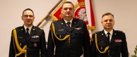 Trzech funkcjonariuszy Państwowej Straży Pożarnej w galowych mundurach pozuje do wspólnego zdjęcia w pomieszczeniu służbowym. Za nimi znajduje się sztandar z godłem Rzeczypospolitej Polskiej oraz flaga państwowa. Funkcjonariusz stojący pośrodku trzyma w dłoni dokument.