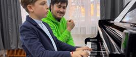 Zdjęcie przedstawiające uczestników warsztatów pianistycznych, które poprowadził mgr Michał Michalski. Przy fortepianie uczeń wraz z prowadzącym podczas lekcji. Warsztaty odbyły się 9 kwietnia w małej auli szkoły, czyli sali numer 3 w "starym" budynku szkoły.