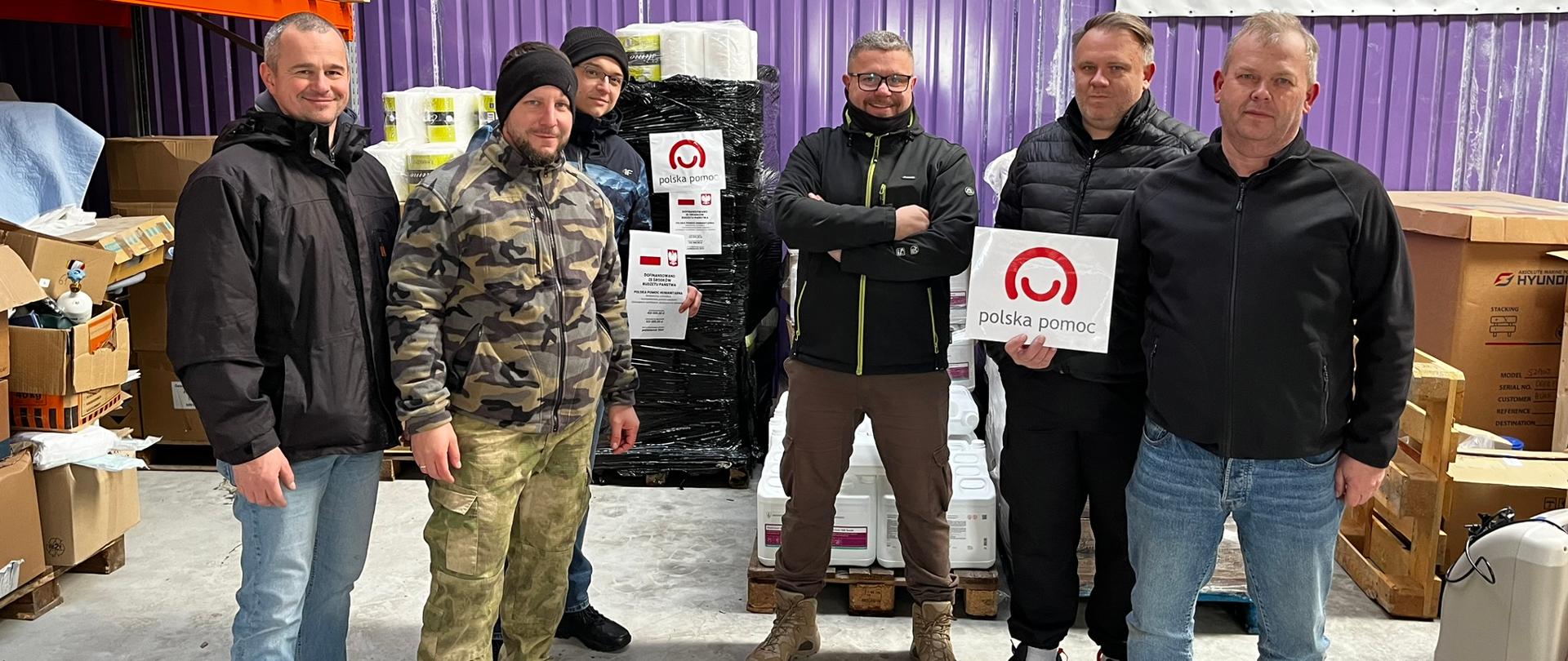 W magazynie na tle pudeł i palet z pomocą materialną do zdjęcia pozuje sześciu mężczyzn 