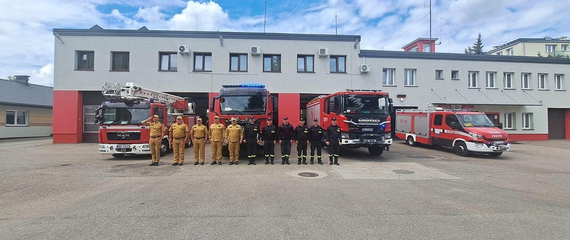 Strażacy stoją w szeregu przed samochodami z włączonymi sygnałami. Jeden z nich salutuje. 