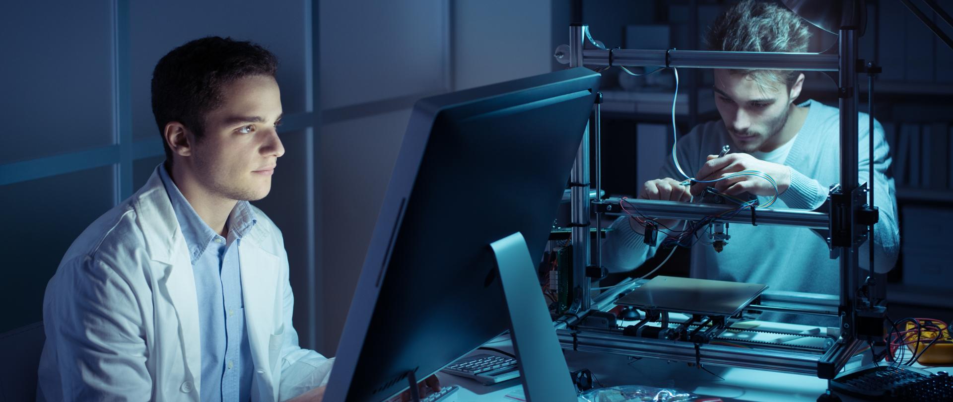 Zdjęcie przedstawia mężczyznę siedzącego przed komputerem i drugiego mężczyznę pracującego w skupieniu nad jakimś technicznym projektem.