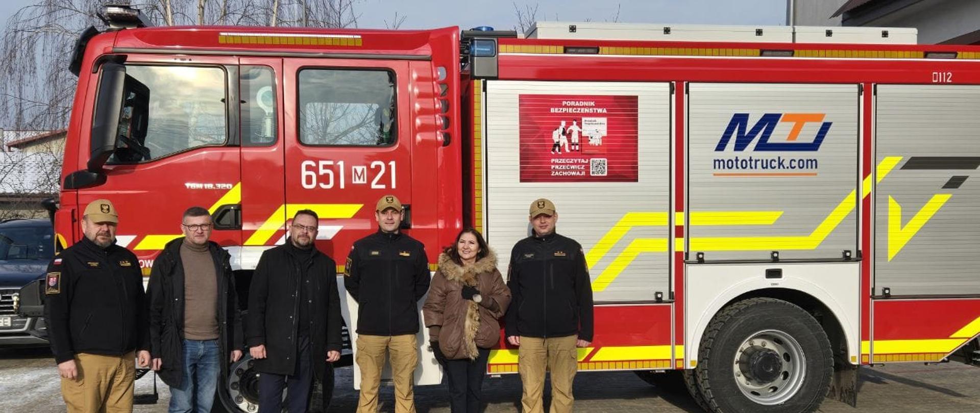 Na zdjęciu grupa sześciu osób. Trzech mężczyzn w ubraniu strażackim, pozostałe osoby to pracownicy starostwa. Za nimi nowy samochód pożarniczy na którym znajduje się naklejka z napisem: ,,Poradnik bezpieczeństwa. Przeczytaj, przećwicz, zachowaj", oraz numery operacyjne pojazdu: 651M21.