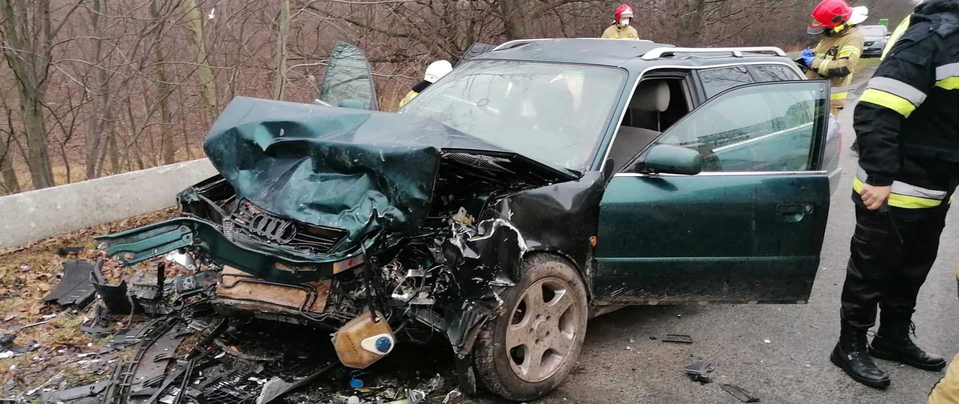 Fotografia przedstawia powypadkowy wrak pojazdu osobowego koloru zielonego. W okół pojazdu widać strażaków prowadzących czynności związane z akcją ratowniczą.