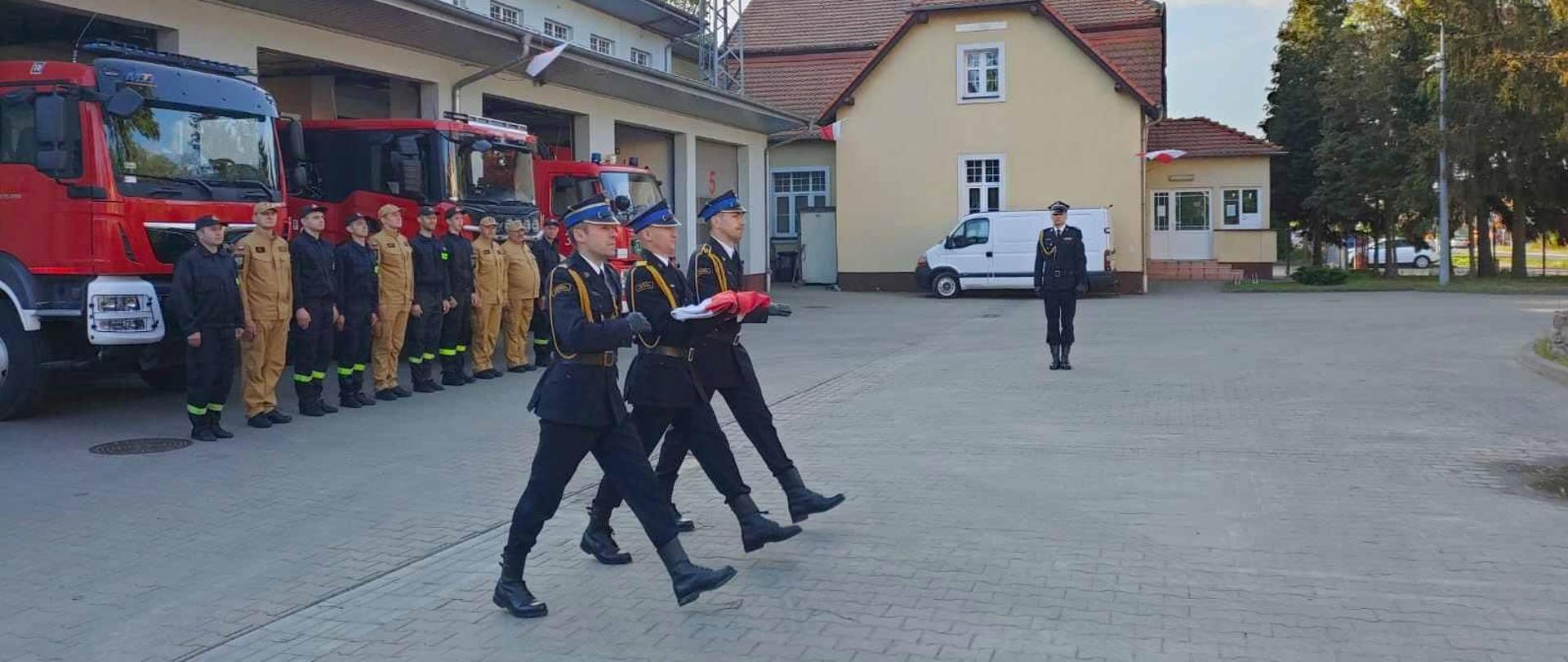 Poczet flagowy niosący flagę, w tle pododdział stojący przed samochodami pożarniczymi i dowódca uroczystości
